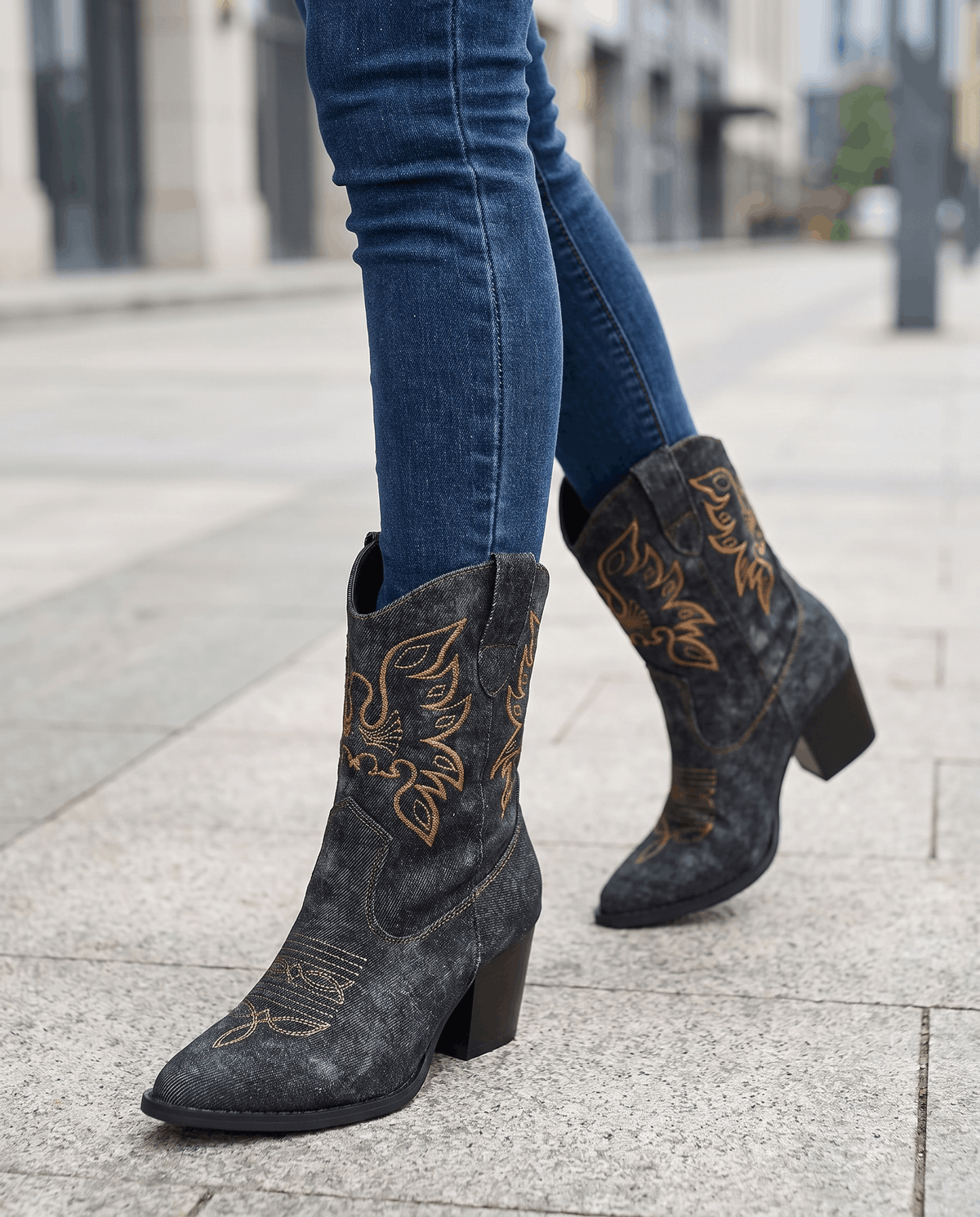 Legs in blue jeans and black embroidered boots with pointed toes on a city sidewalk
