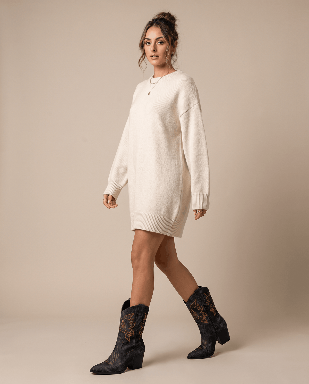 Woman in cream sweater dress walking in black embroidered boots against a tan backdrop
