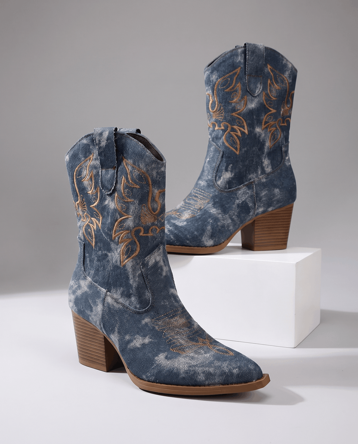 Two blue embroidered boots with brown block heels displayed on a white block