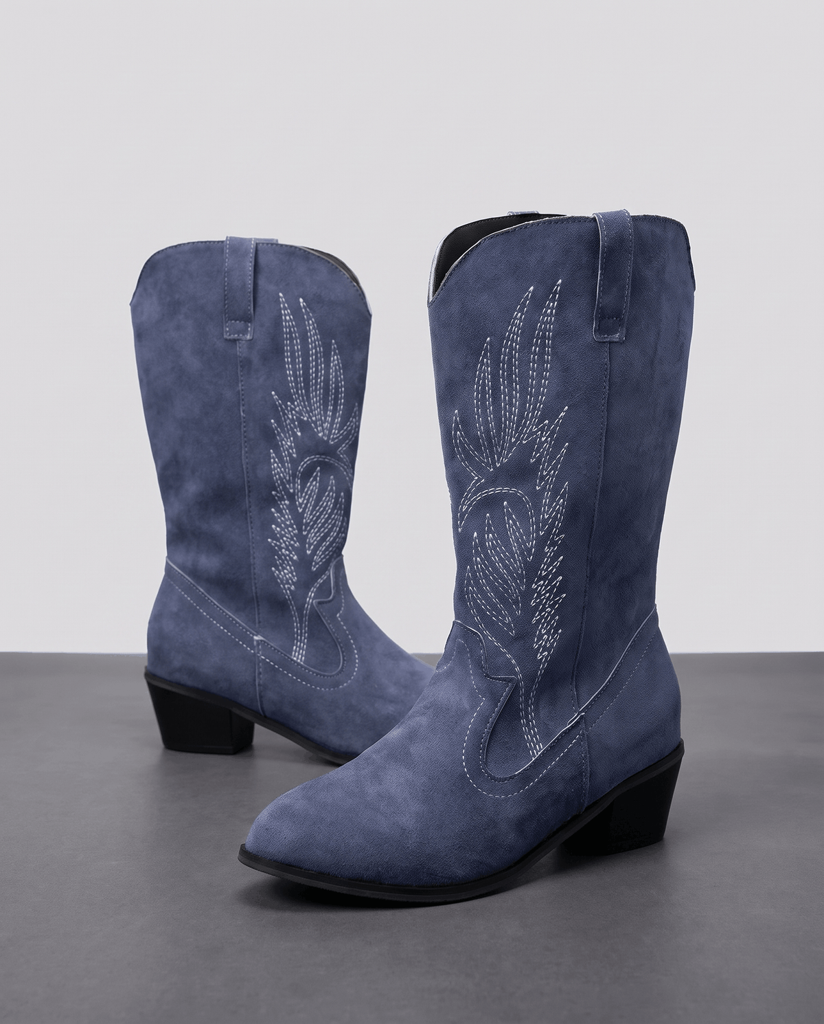 Two navy blue suede boots with light embroidery and black block heels against a grey backdrop
