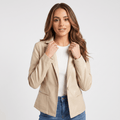 Woman wearing a beige faux leather blazer with a notched lapel, single-button closure, front flap pockets, and a tailored fit over a white top and blue jeans.