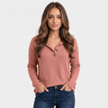 Woman wearing a dusty rose ribbed henley sweater with a button neckline, long sleeves, and fitted silhouette with blue jeans.