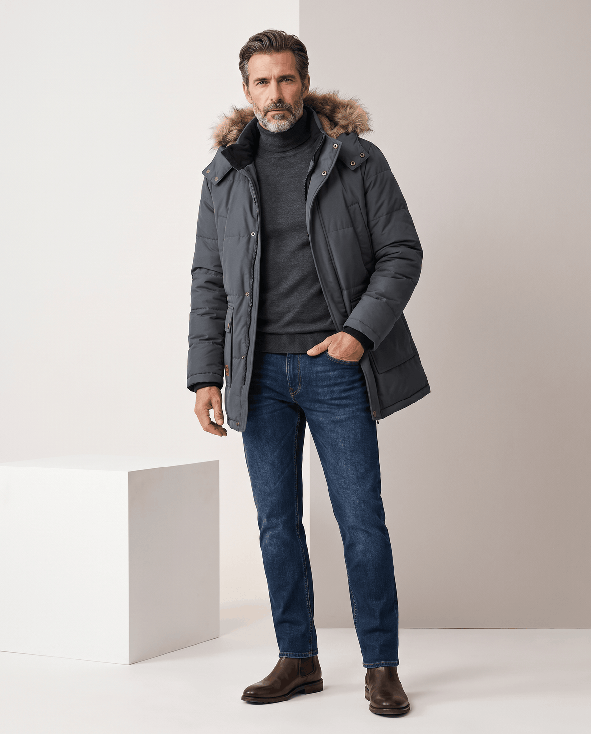 Man in dark blue parka, dark jeans and brown shoes standing with one hand in pocket