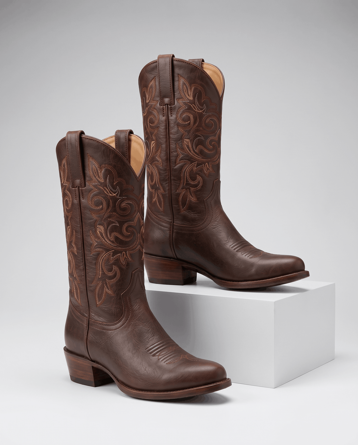 Two brown boots with tonal embroidery and pull tabs, one resting on a white block