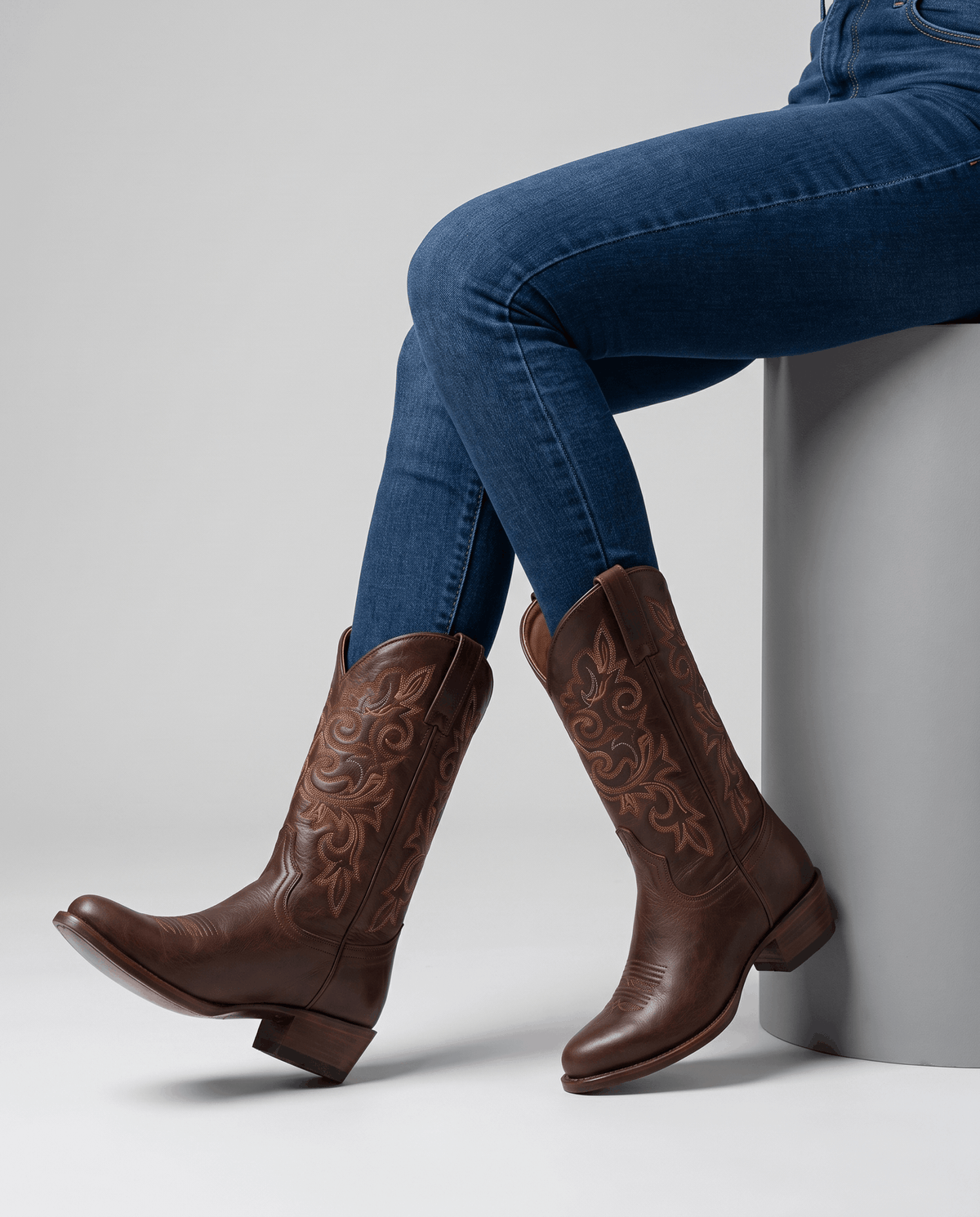 Seated legs in blue jeans and brown embroidered boots against a grey studio backdrop