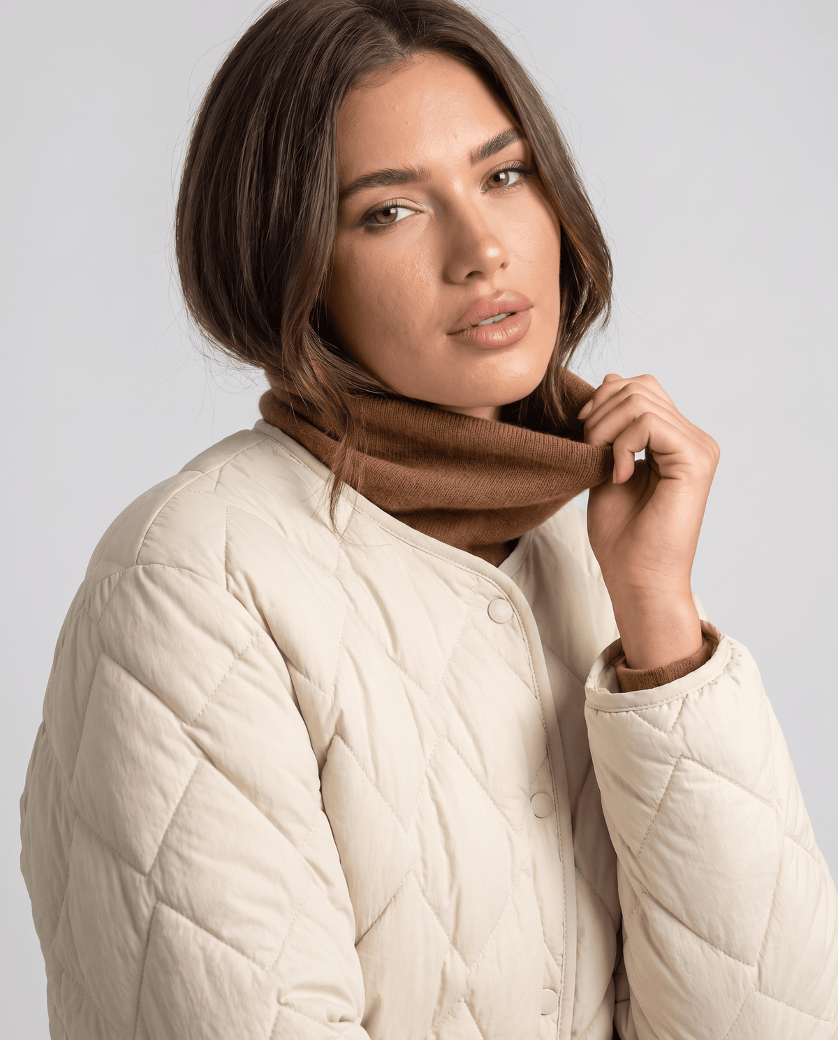 Model in cream quilted jacket pulling down brown turtleneck, close-up of diamond stitching