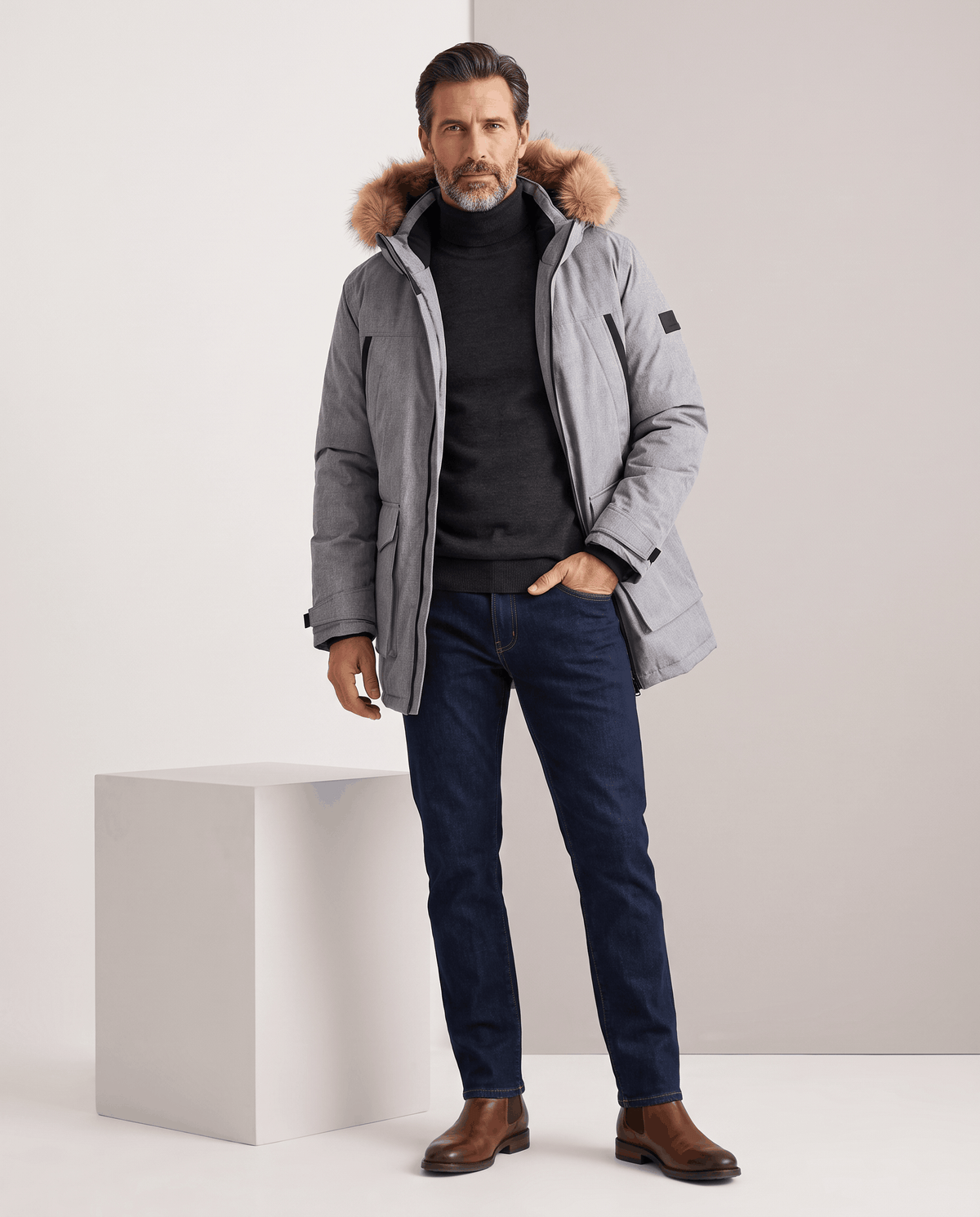 Man in gray parka, dark jeans and brown boots with one hand in pocket
