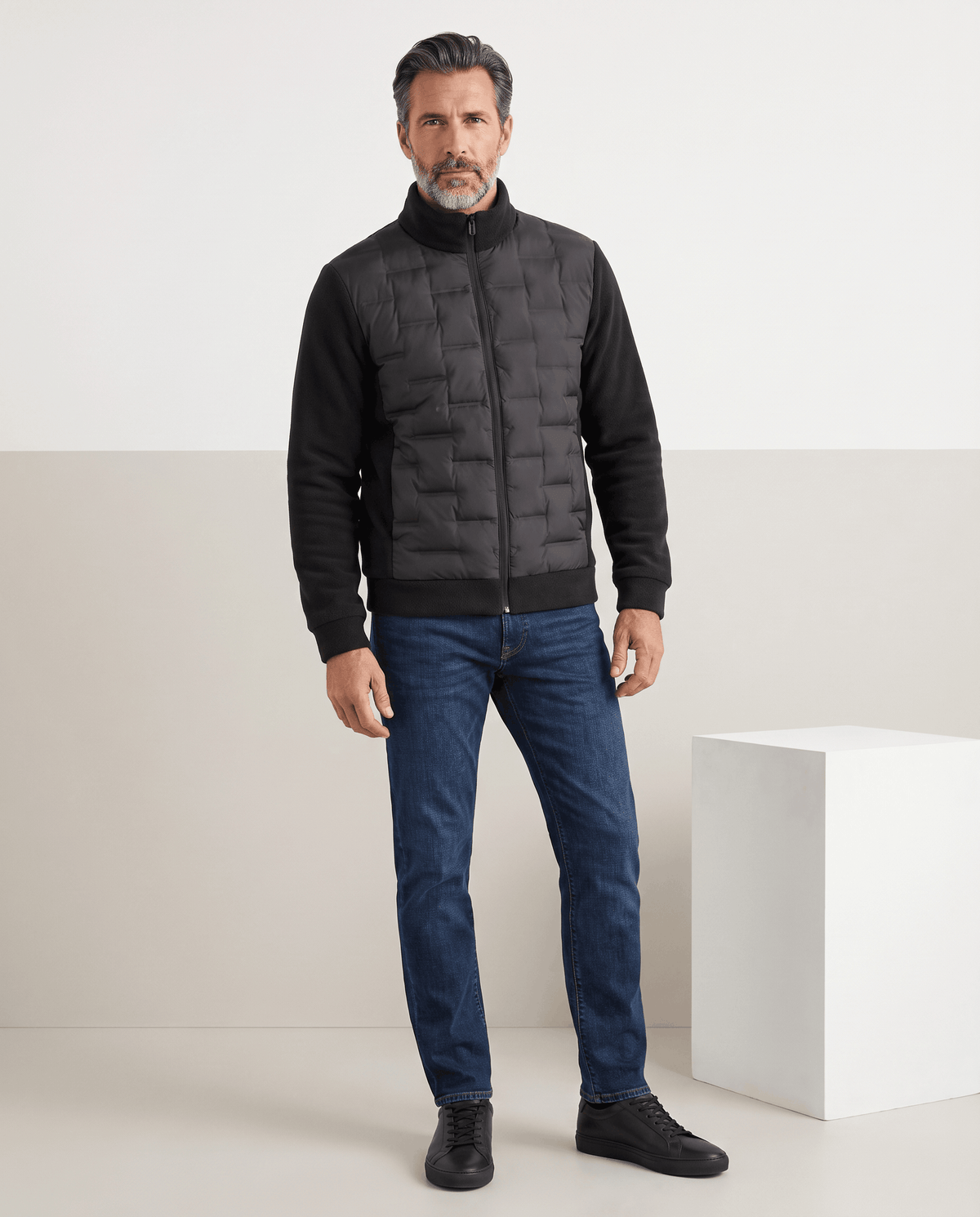Man standing in a black hybrid puffer jacket with blue jeans, full-length view