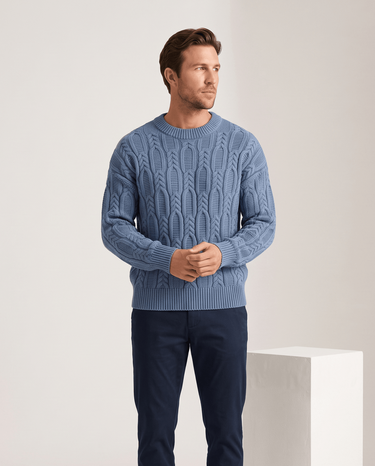 Model in blue crew neck cable sweater with navy trousers beside a plinth
