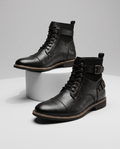 Black leather cap-toe lace-up boots featuring layered panels, adjustable buckle straps, and a welted stacked heel.