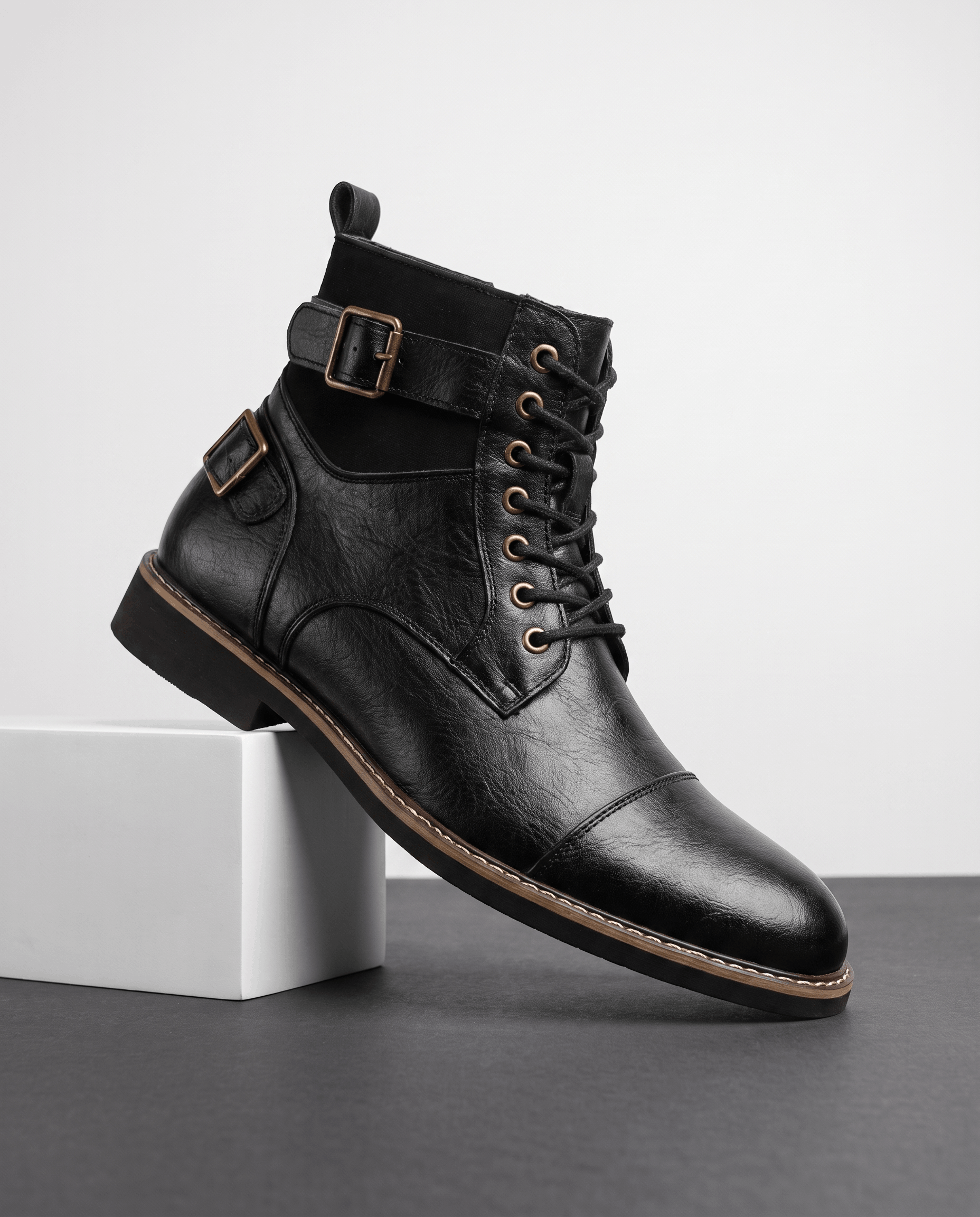Black leather cap-toe lace-up boot with layered panels, adjustable buckle straps, and a welted stacked heel.