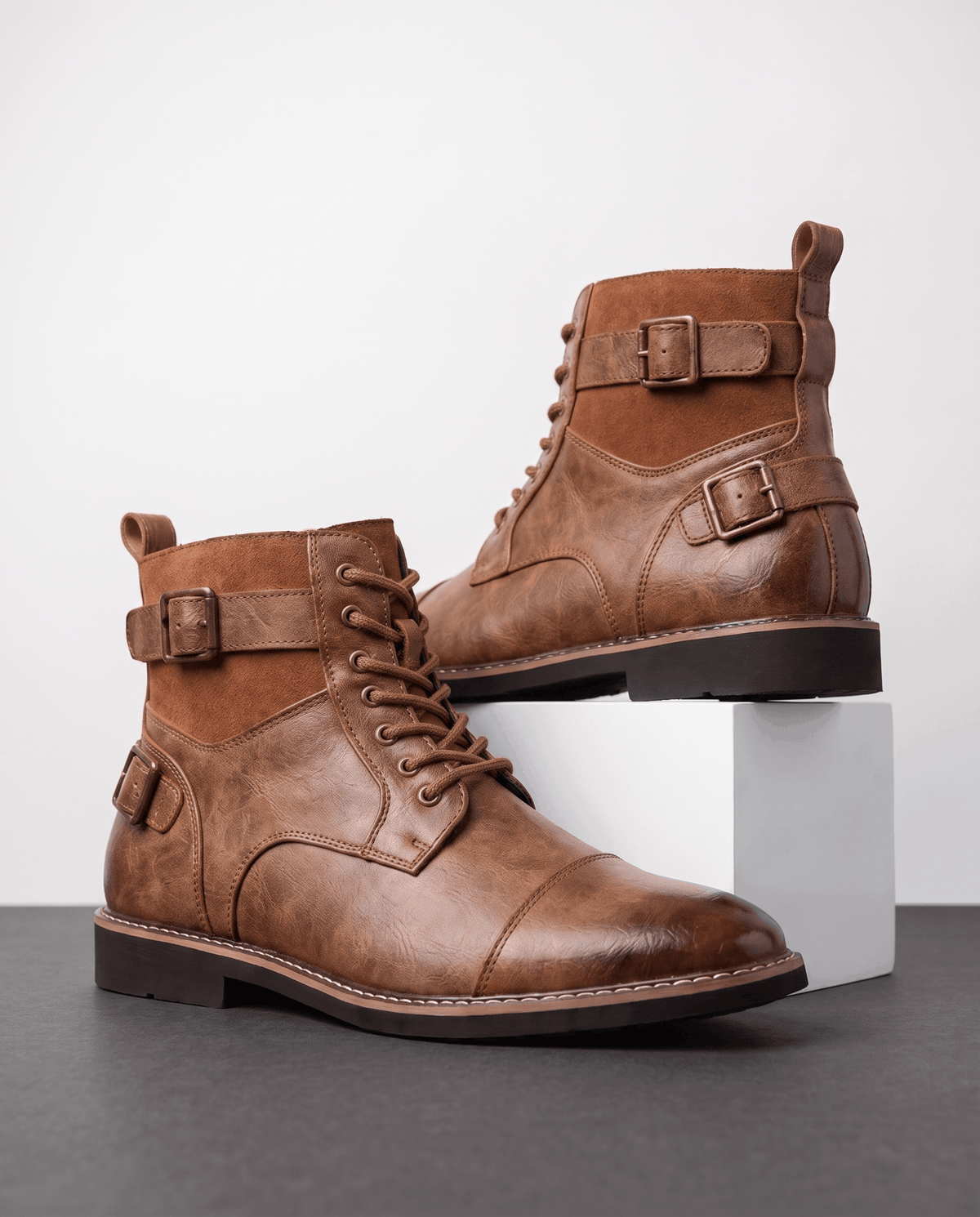 Brown cap-toe lace-up boots featuring layered panels, dual adjustable buckle straps, and a stacked welted heel.