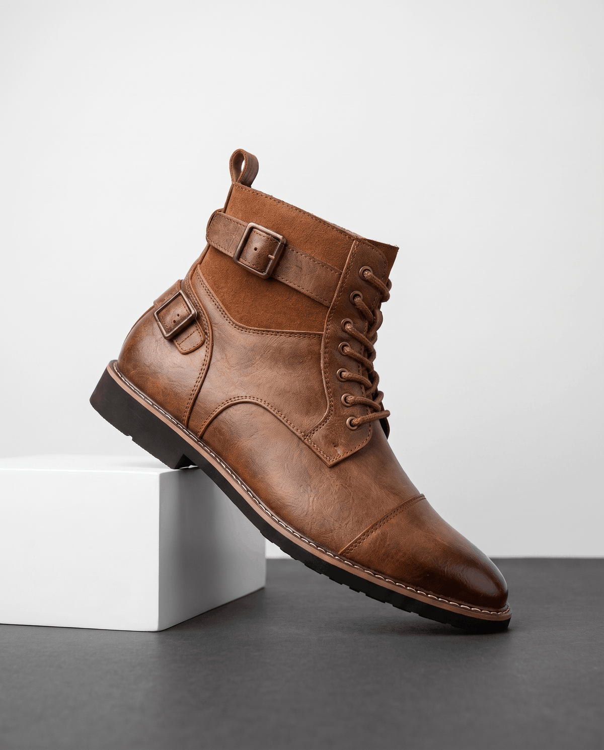 Brown cap-toe lace-up boot featuring layered leather panels, a suede collar, and an adjustable side buckle strap.