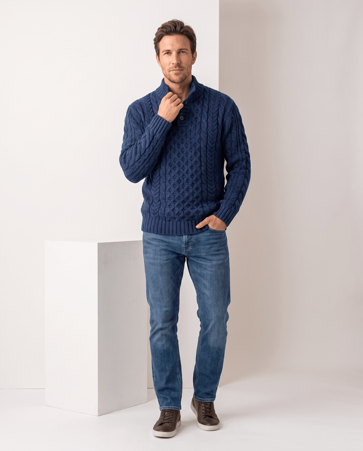 Full-length navy cable sweater outfit with jeans beside a white plinth