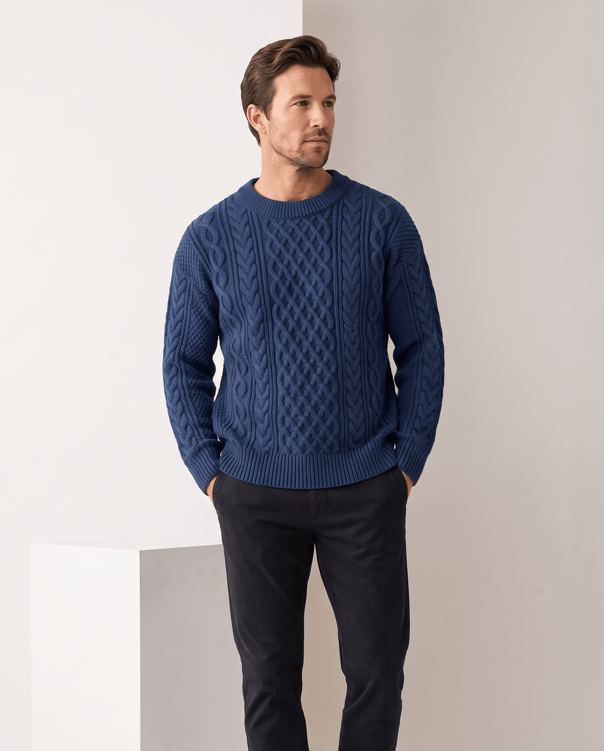 Model in blue crew neck cable sweater with black trousers, hands in pockets