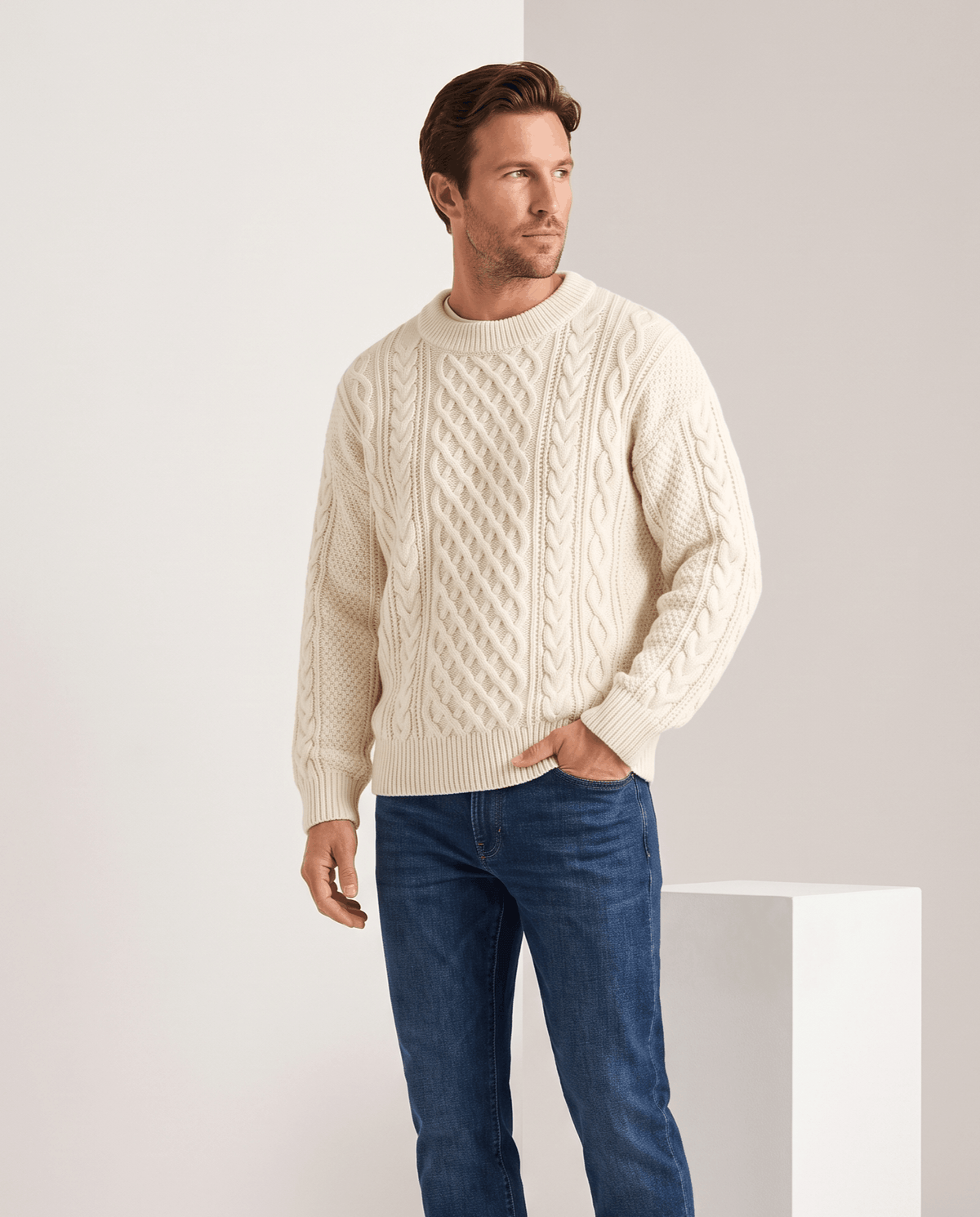 Model in cream cable sweater with blue jeans beside a white plinth