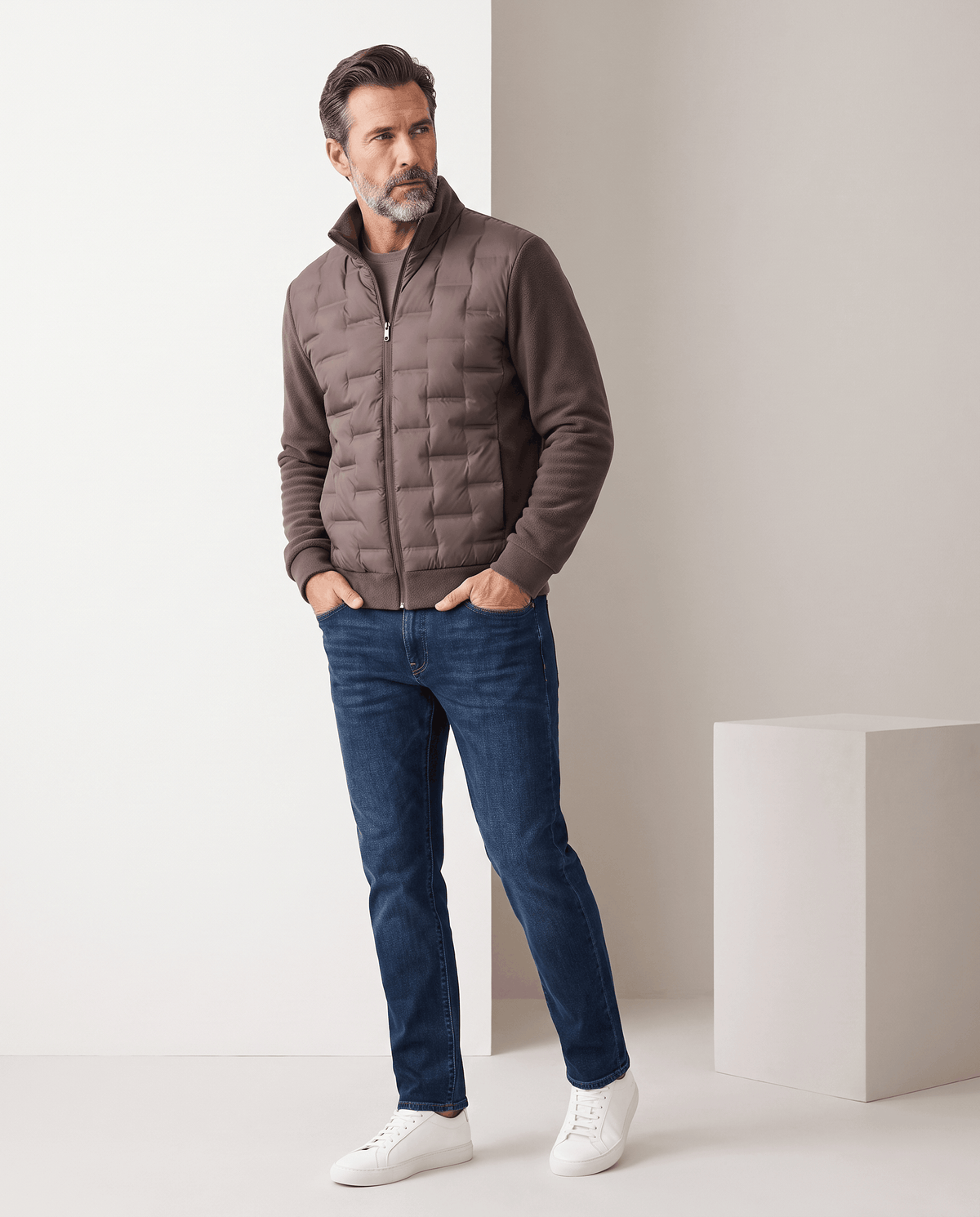 Man standing in a dark brown hybrid puffer jacket with blue jeans, full-length view