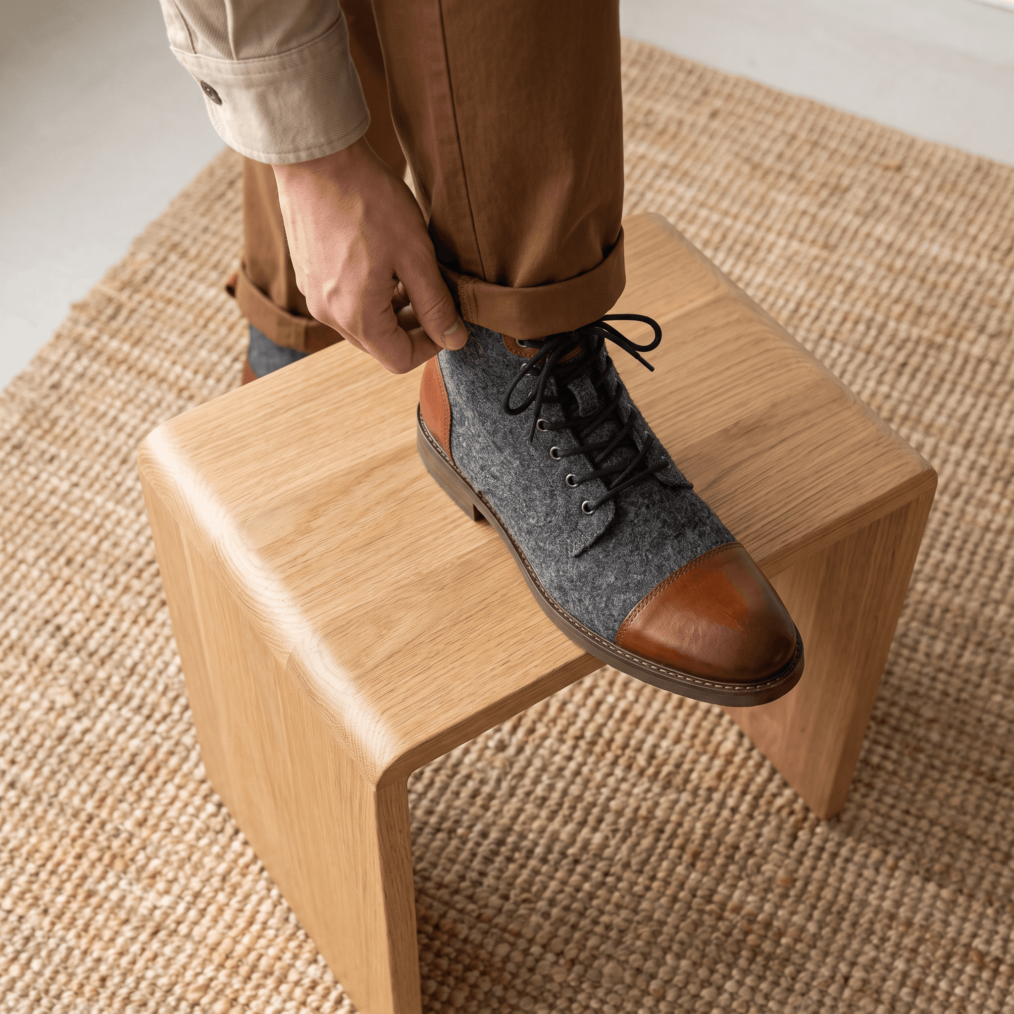 Brown felted wool and brown leather cap toe lace-up boots with a welted sole, worn with brown trousers on a wooden stool.