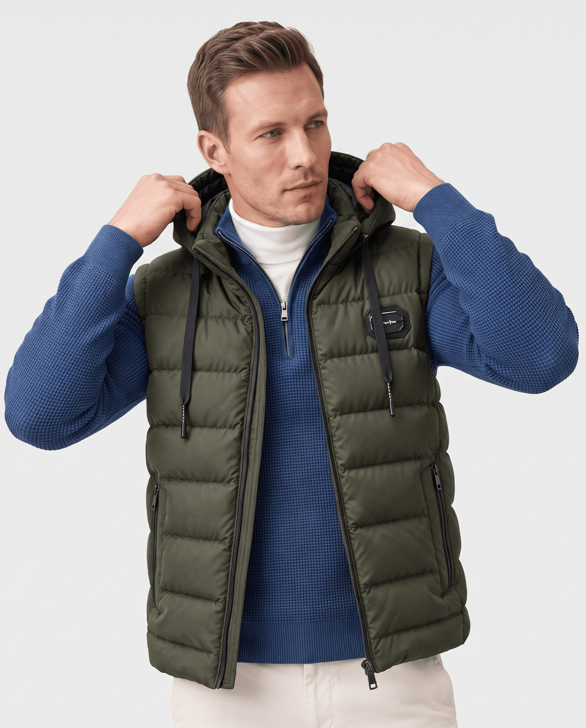 Men's hooded puffer vest in green worn over a blue zip-neck sweater, hands at the hood