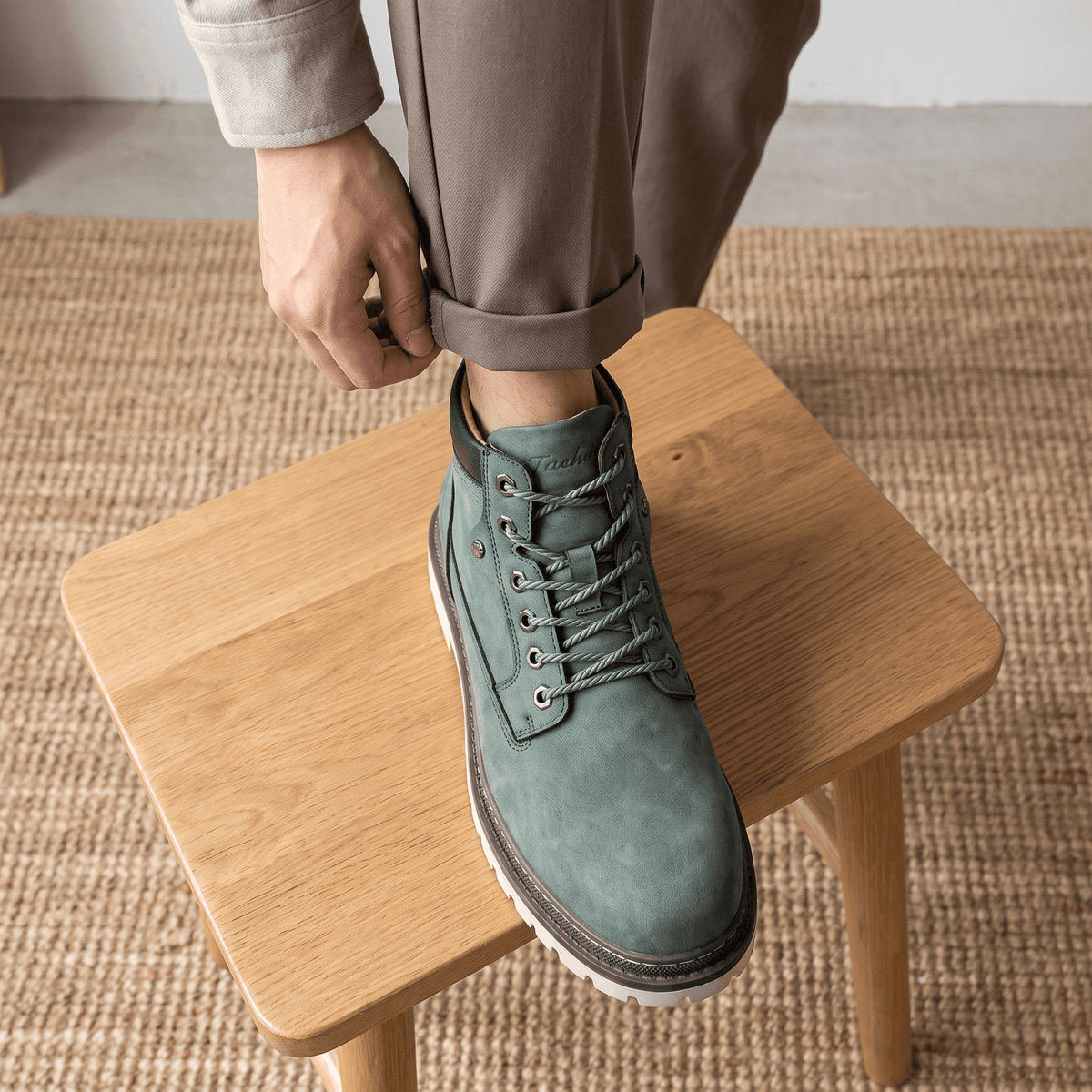 Matte sage green lace-up work boots featuring a padded collar, bolt-style eyelets, and a chunky lug sole on a wooden stool.