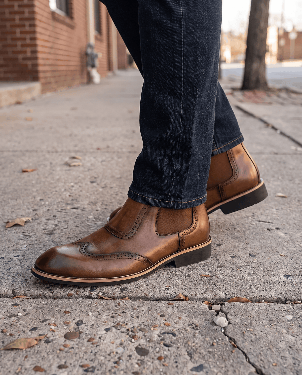Man in tan brogue slip-on boots and dark trousers walking along a sidewalk