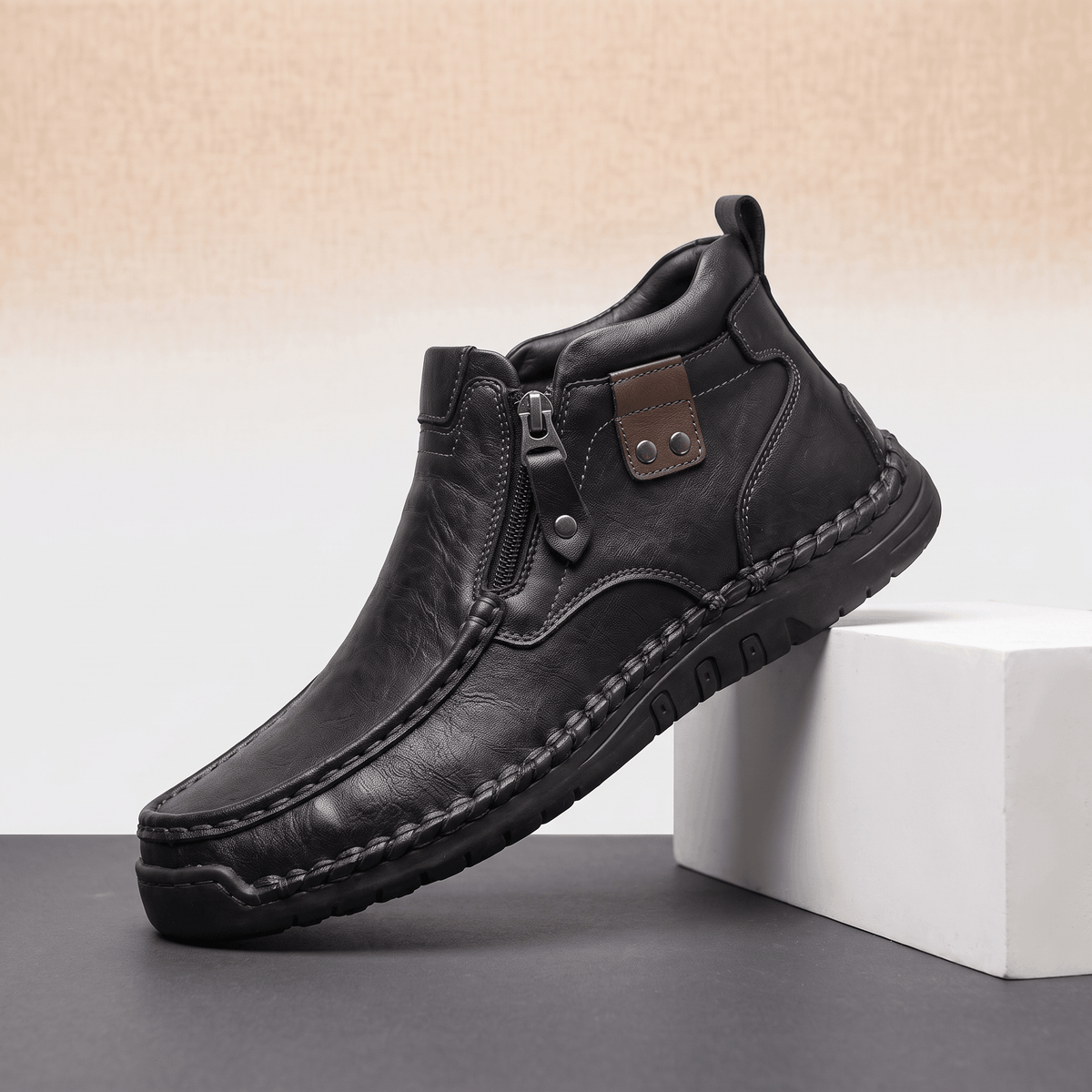 Black leather moc toe boot with side zip, apron seam, and visible whipstitched rubber midsole on a white display block.