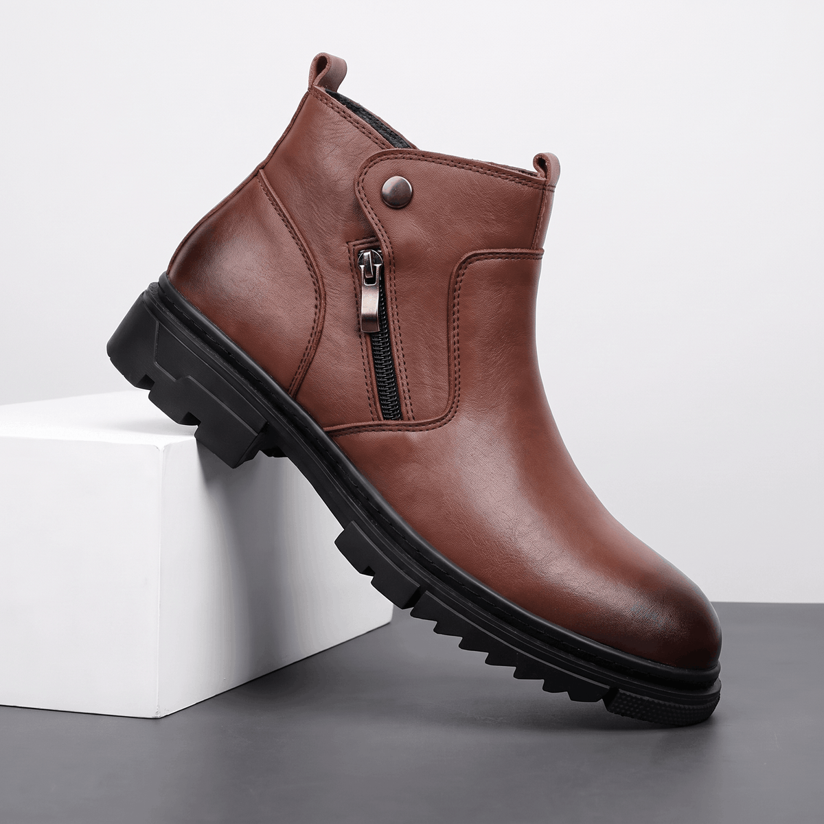 Smooth brown leather ankle boot with a side zip, snap tab detail, and a chunky black lug sole on a white block.