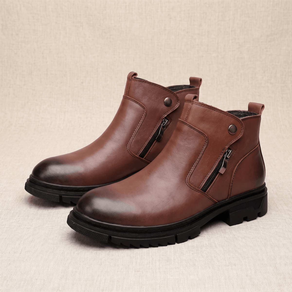 Smooth brown leather ankle boots with side zip closures, snap tabs, and chunky black lug outsoles on a textured background.