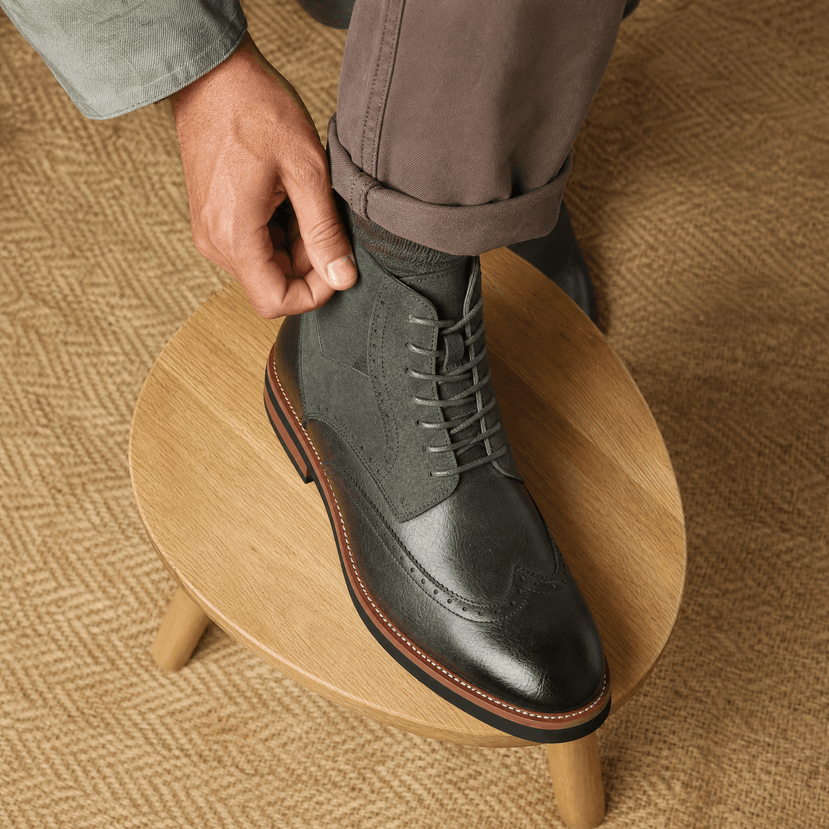 Gray leather wingtip ankle boots featuring a suede shaft panel, burnished toe, and welted rubber sole on a wooden stool.