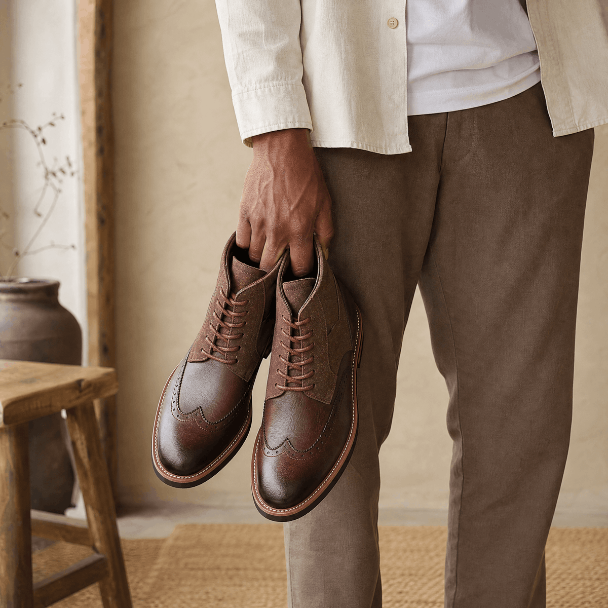 Dark brown leather wingtip ankle boots with suede shafts, burnished toes, and welted rubber soles held by a person.
