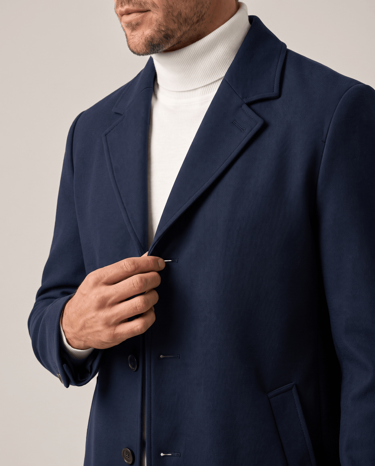 Hand on the lapel of the navy coat, with a white turtleneck underneath and a slant pocket below