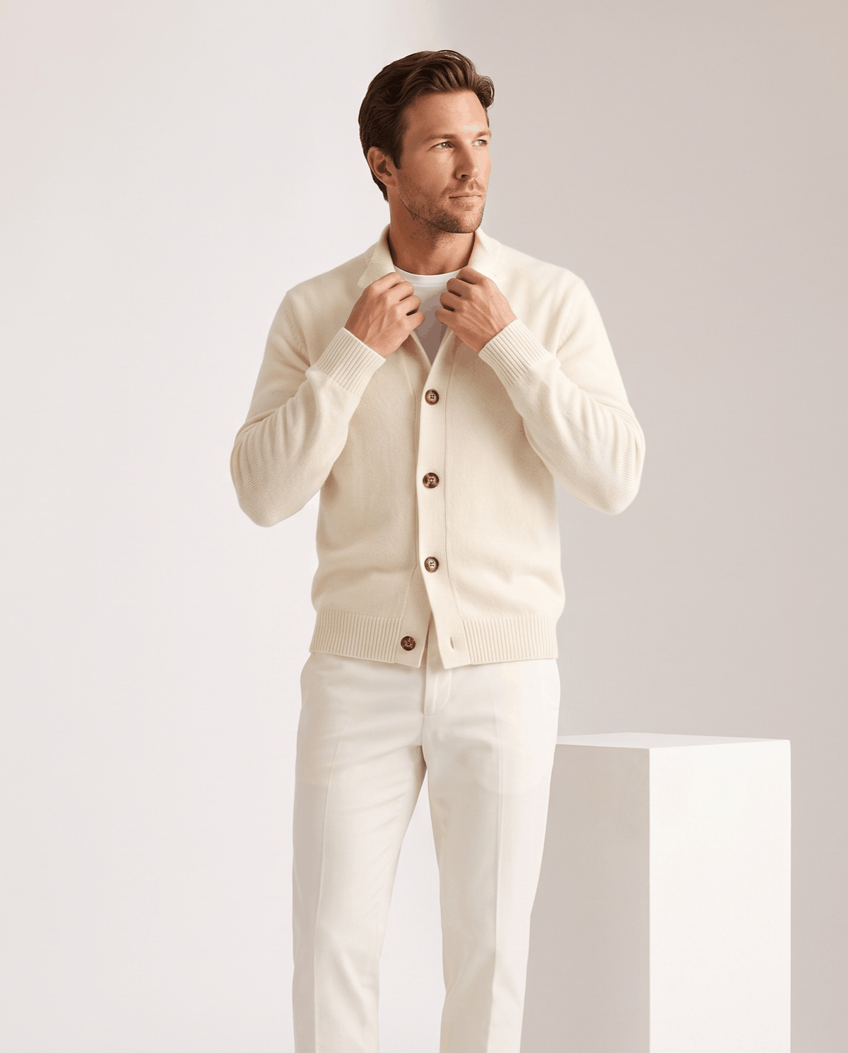 Man buttoning an ivory knit cardigan worn with white trousers