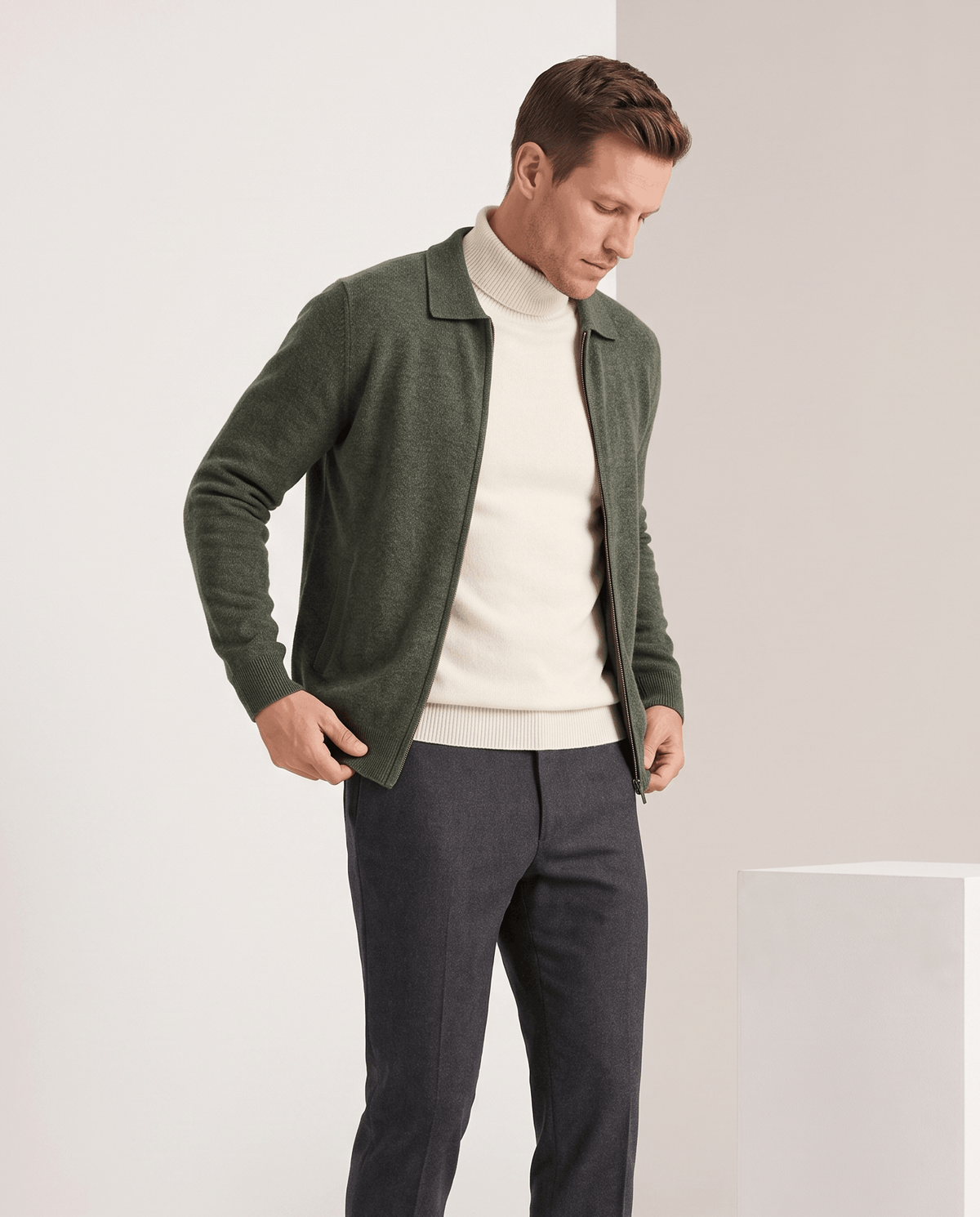 Olive green zip cardigan worn open over a cream turtleneck with charcoal trousers