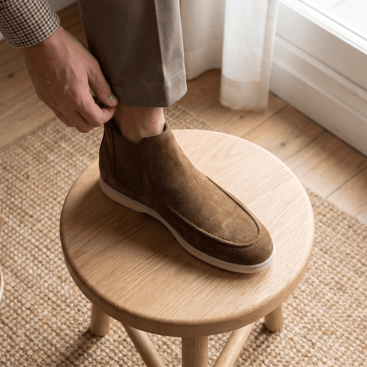 Person putting on a brown suede moc toe Chelsea boot while seated on a wooden stool.