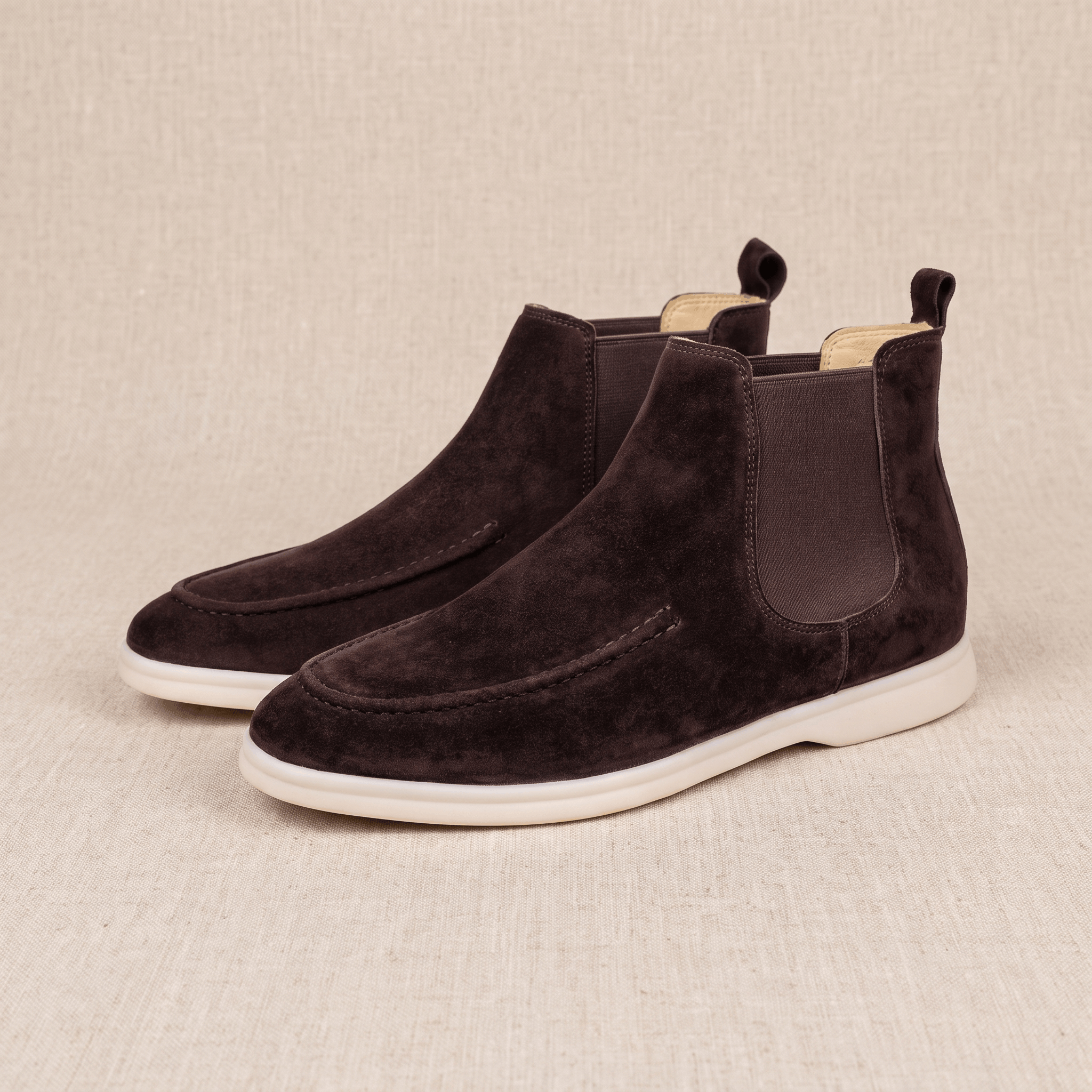 Pair of dark brown suede moc toe Chelsea boots with elastic side panels, pull tabs, and light cupsoles.