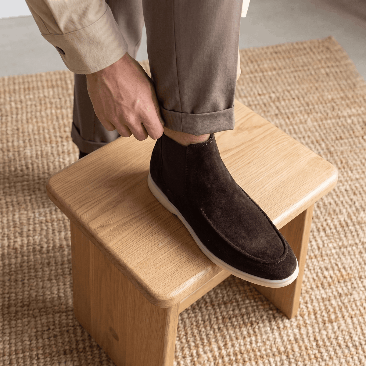 Person putting on a dark brown suede moc toe Chelsea boot while seated on a wooden stool.