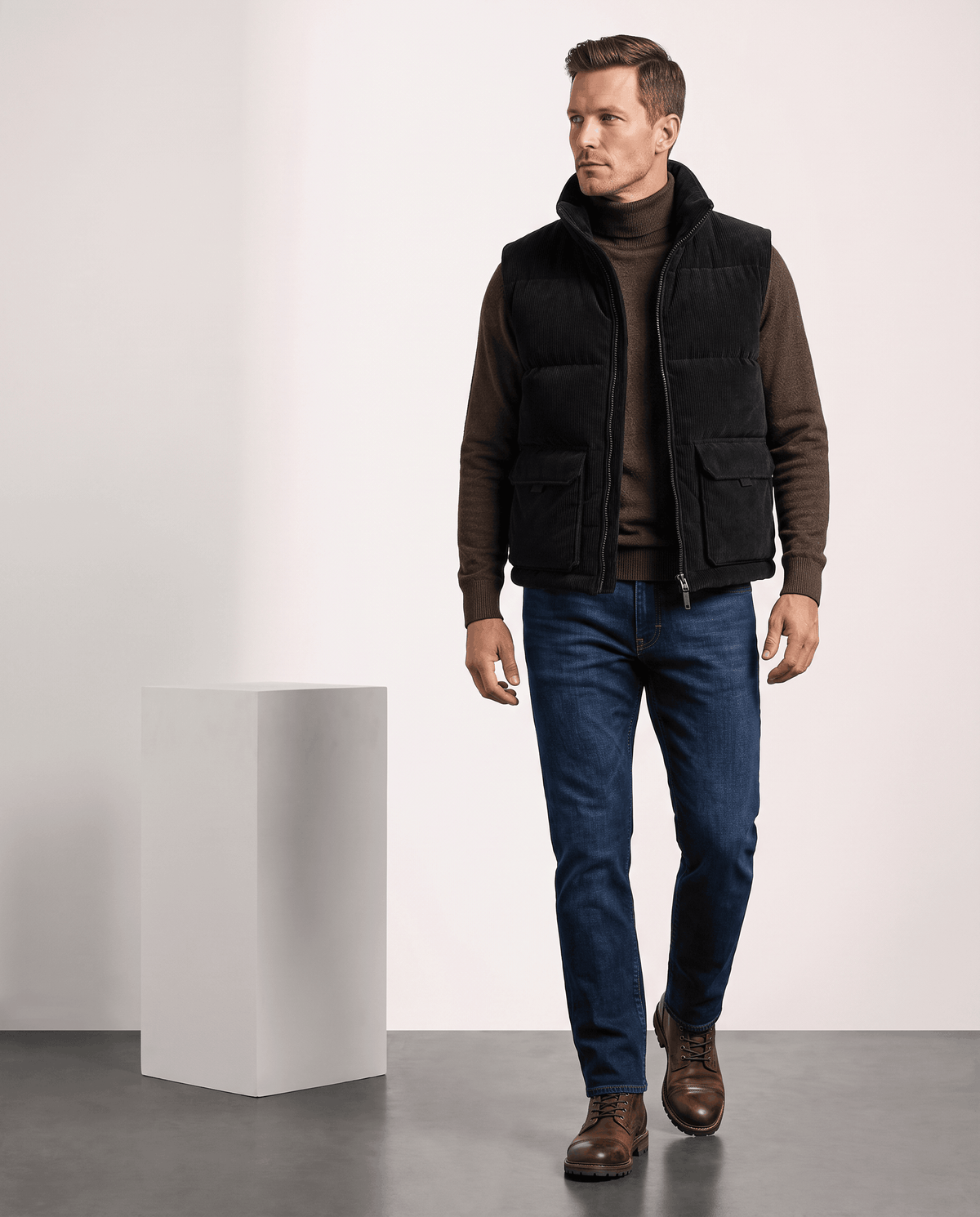 Man in black corduroy vest over a brown sweater with jeans, full length in studio