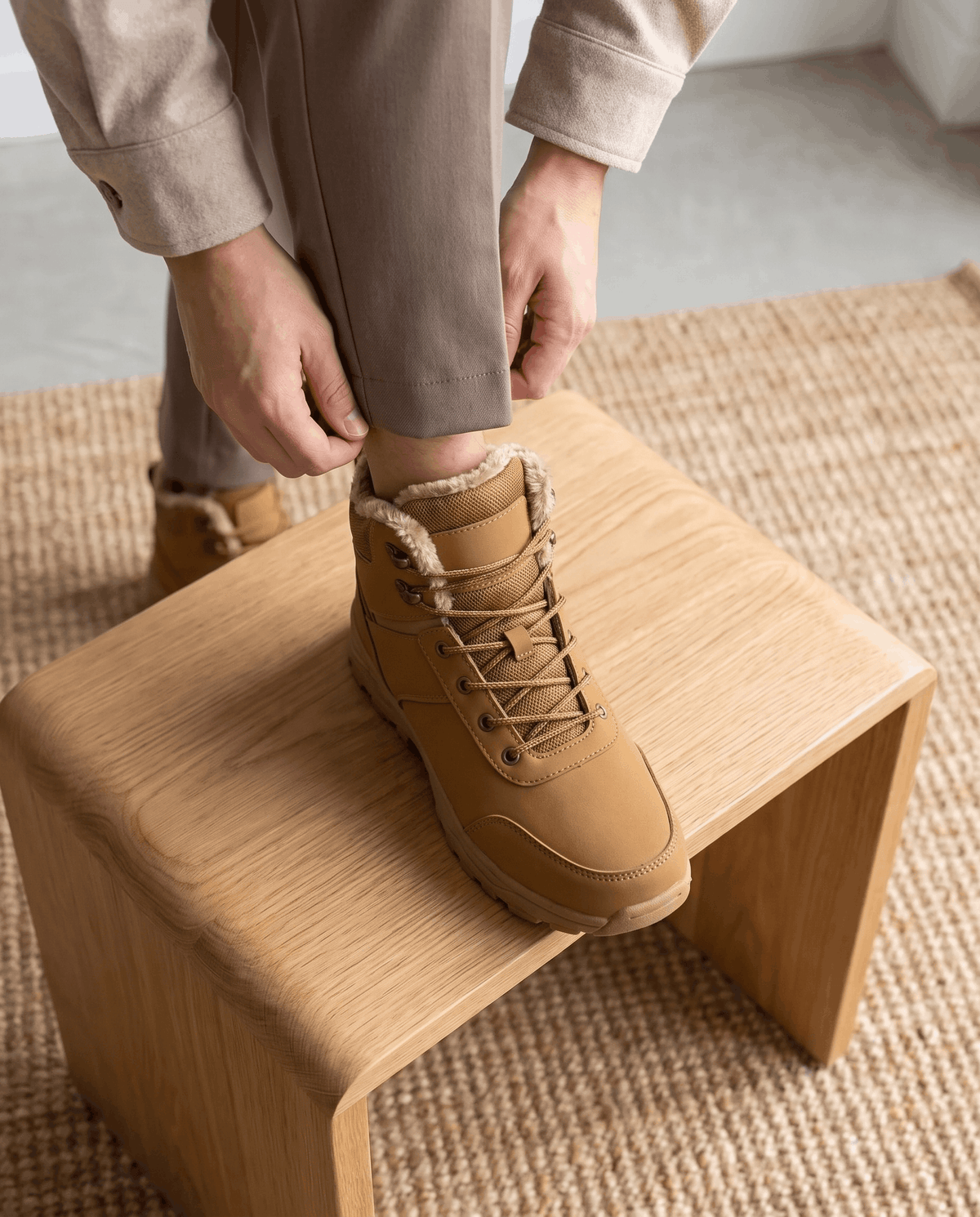 Person putting on a tan winter boot while seated on a wooden stool.