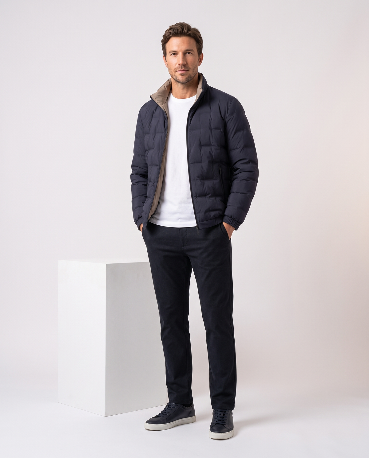 Man wearing the blue Sutton quilted down jacket zipped, standing beside a white plinth