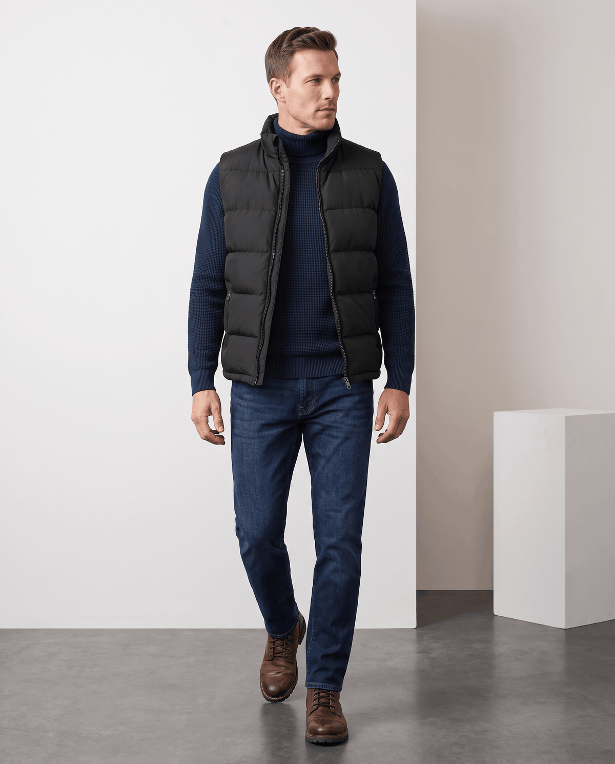 Man in black quilted vest over a dark sweater with jeans and brown shoes, full length