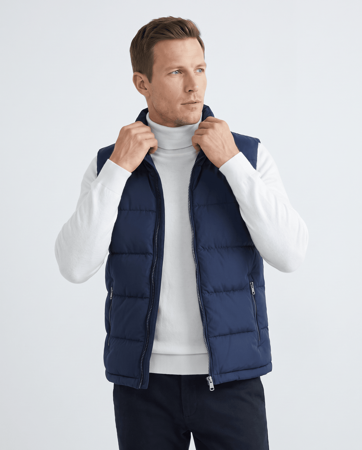 Men's quilted puffer vest in navy worn over a white turtleneck, hands at the collar