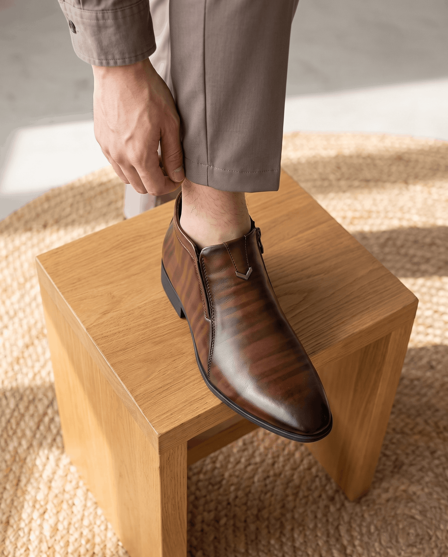 Polished brown grain leather ankle boots with a pointed toe, side zip closure, and notched topline worn with grey trousers.