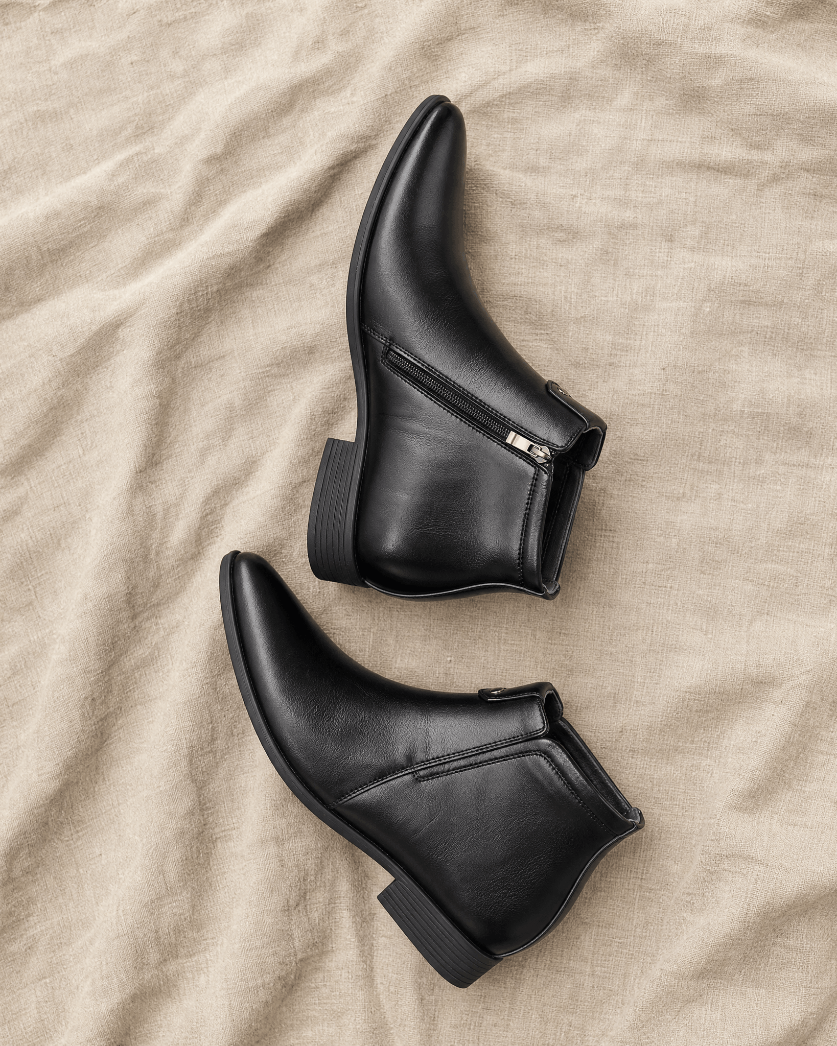 Black polished grain leather ankle boots with pointed toes, side zip closures, and low block heels on a textured beige fabric.
