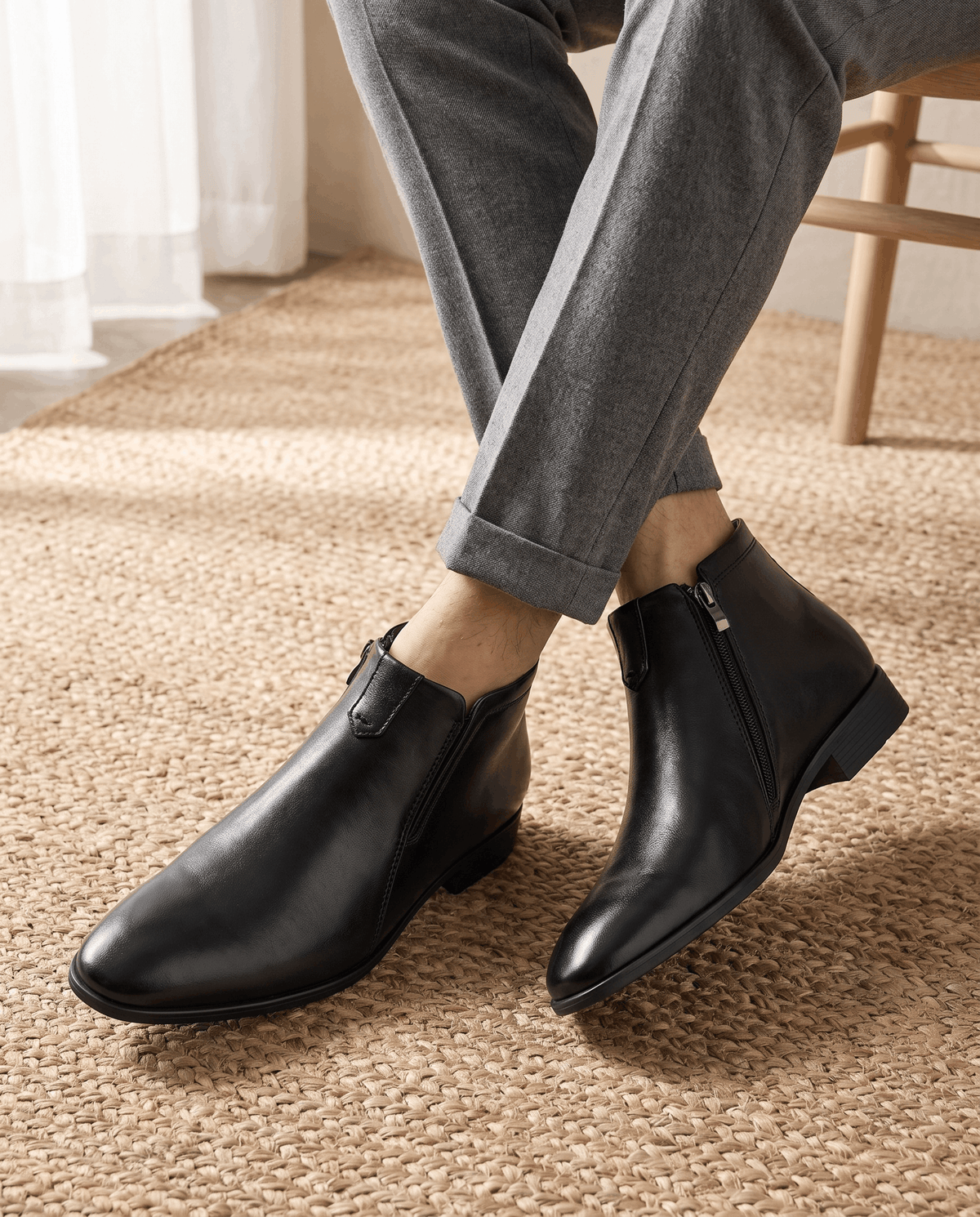 Black polished grain leather ankle boots with a pointed toe, side zip closure, and notched topline worn with grey trousers.