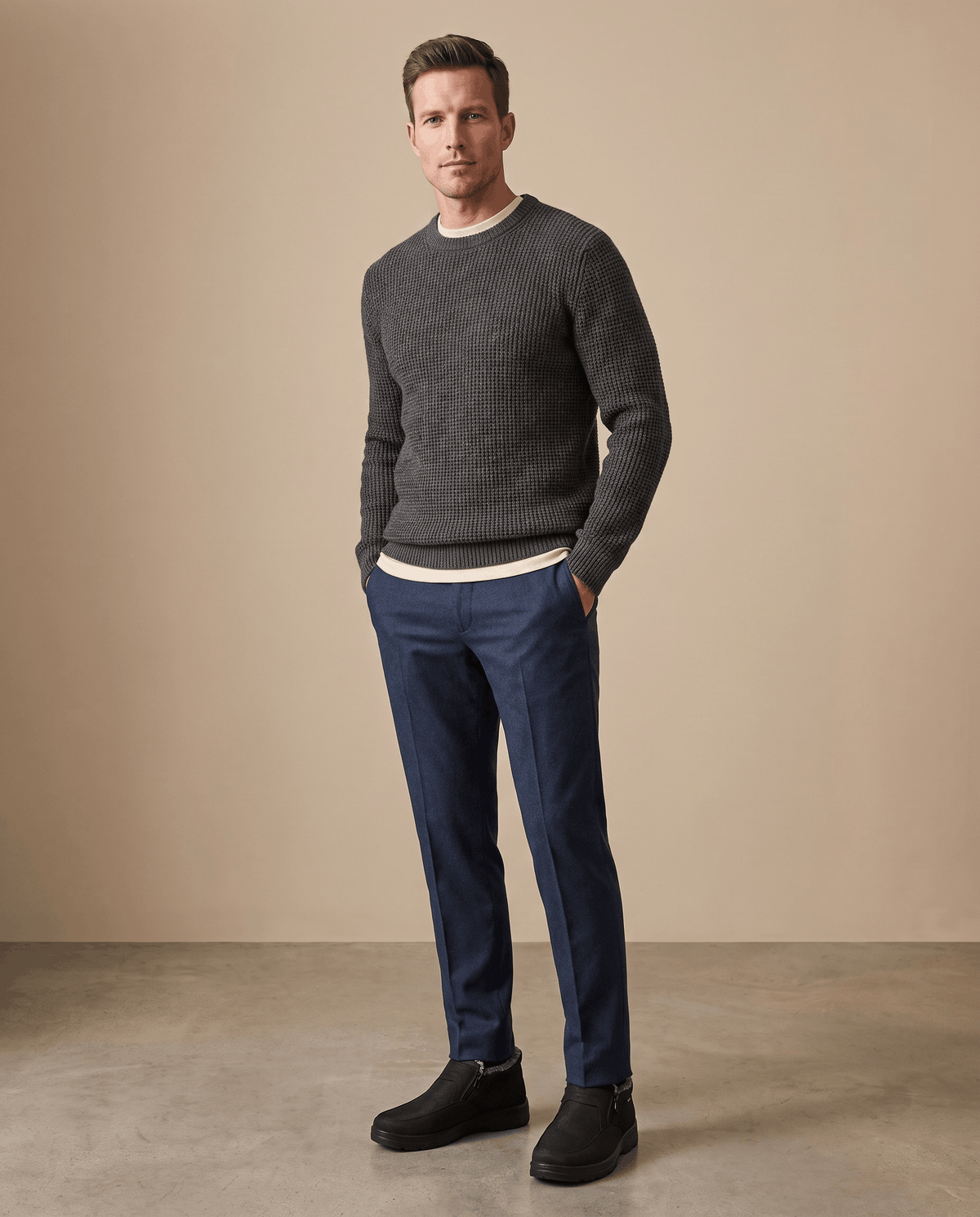 Man wearing black ankle boots with dark pants and a sweater in a minimalist setting.