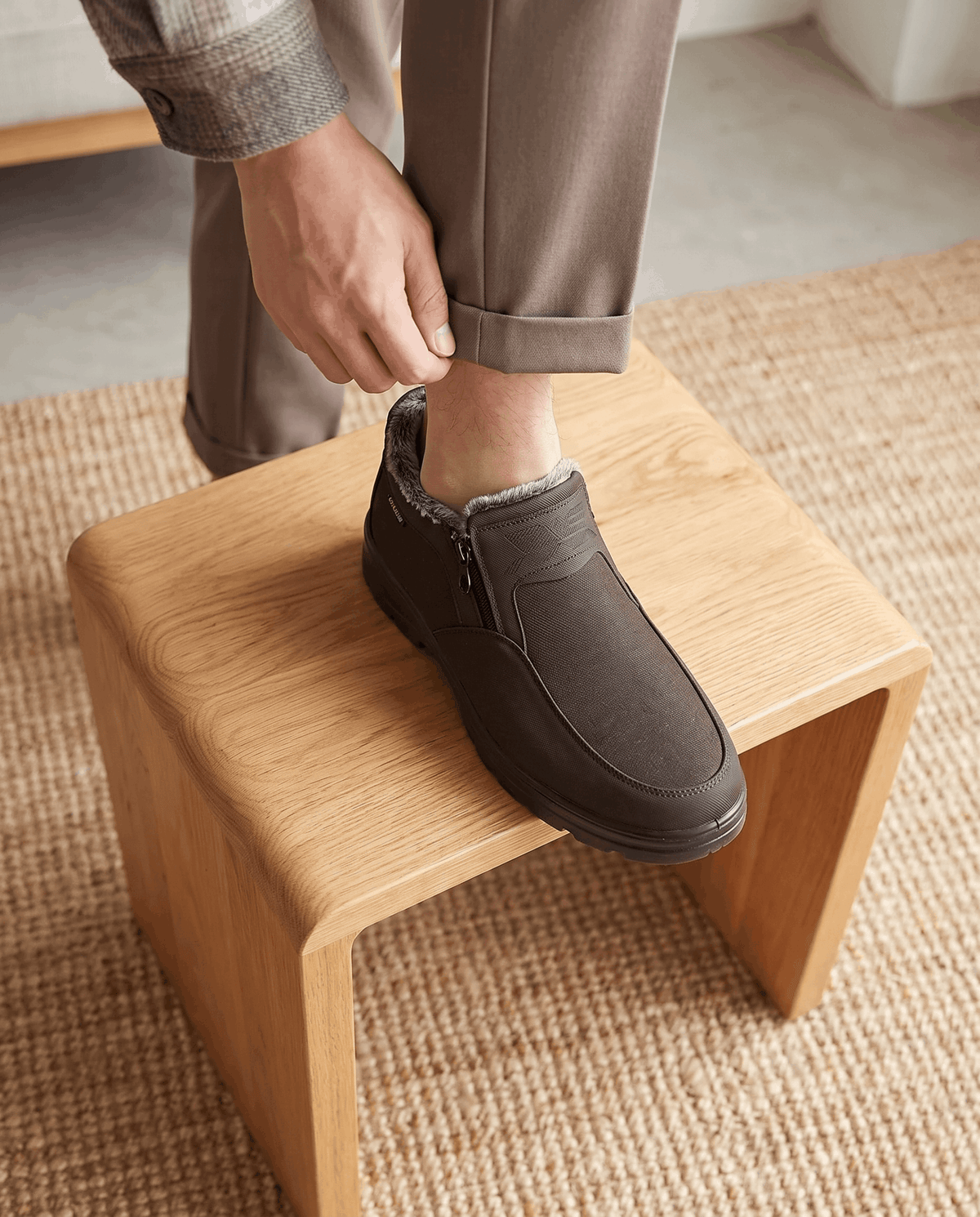 Person putting on a brown fleece-lined ankle boot while seated on a wooden stool.