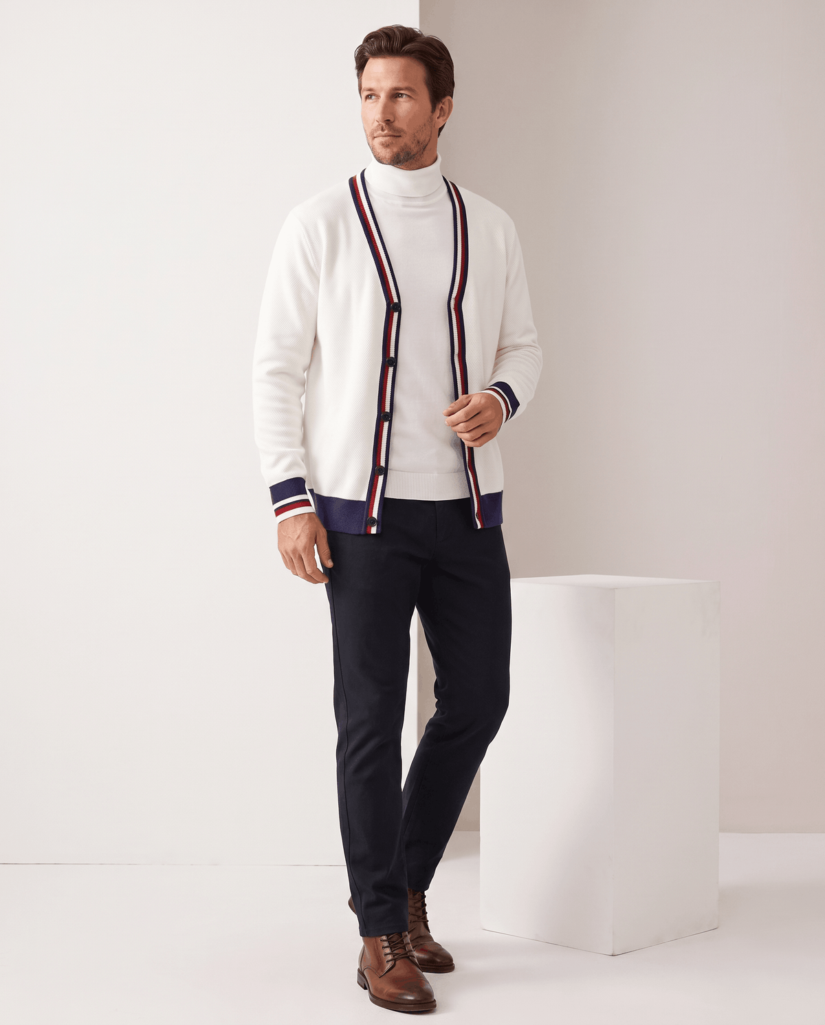 Man in a cream white stripe-trim V-neck cardigan sweater with dark jeans and brown shoes