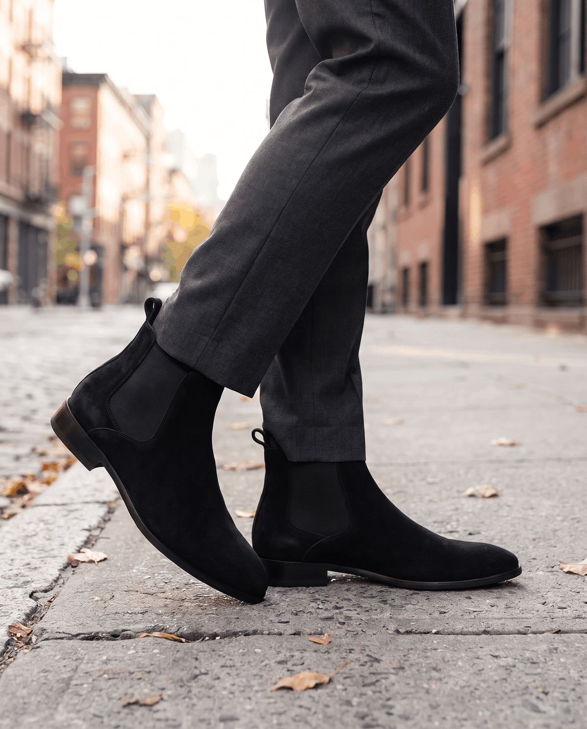 Man in black suede Chelsea boots and gray trousers walking on a city sidewalk