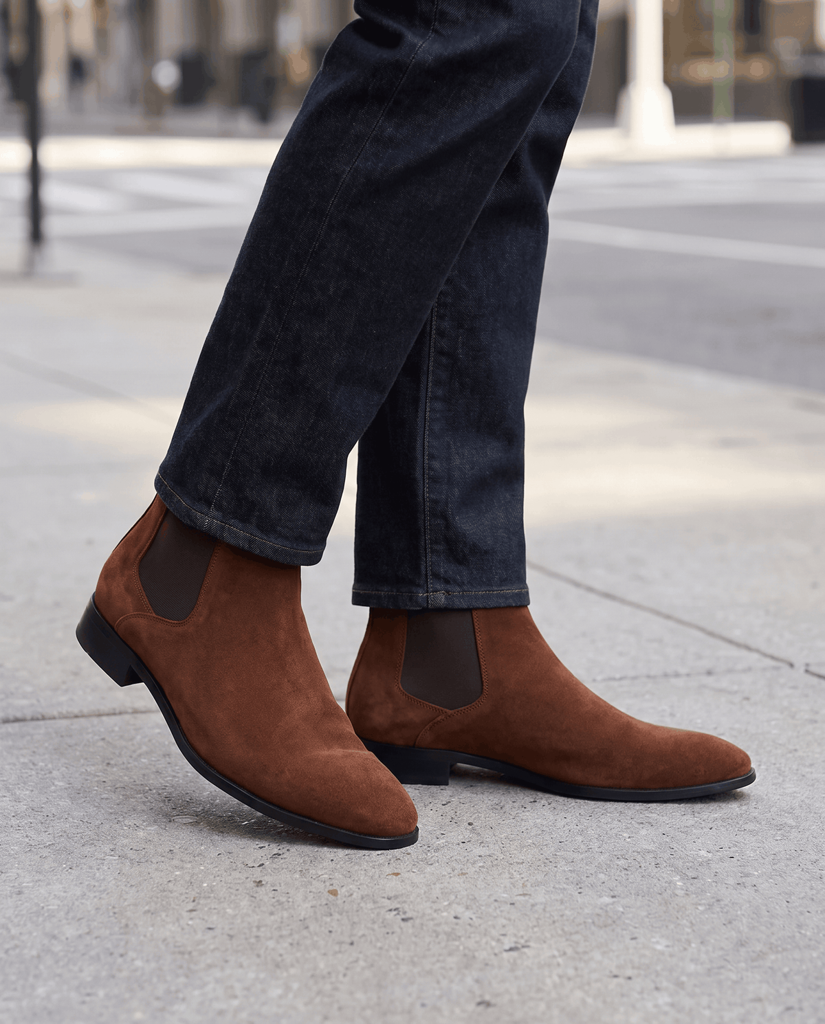 Man in rolled blue jeans and brown suede Chelsea boots stepping on a paved street