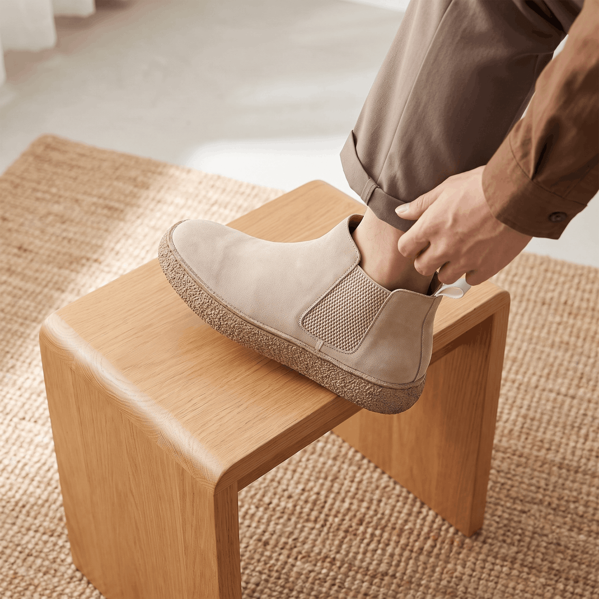 Person putting on a beige suede Chelsea boot while seated on a wooden stool.